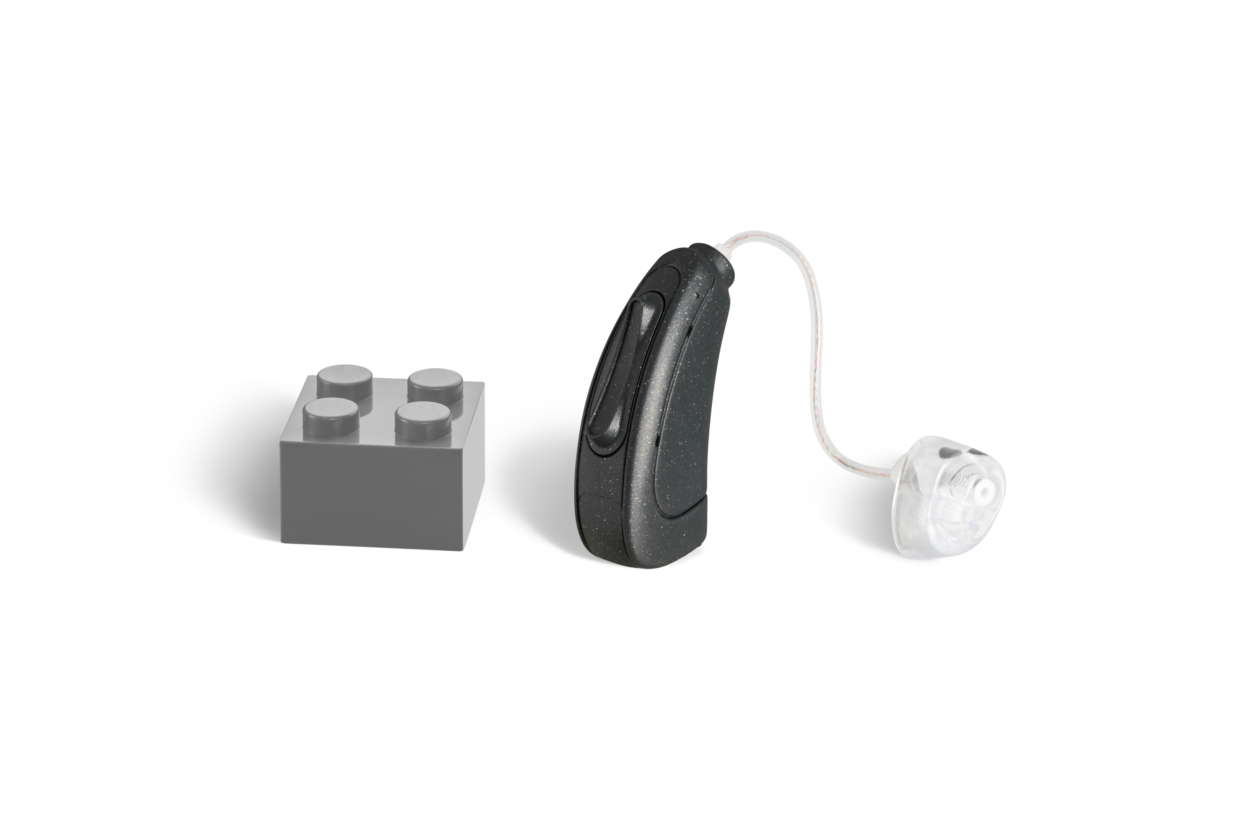 Das Hörgerät KINDaluna 1400 als Bauform HdO mit externem Hörer mit Energieversorgung Batterie neben einem grauen Klemmbaustein stehend auf weißem Hintergrund.