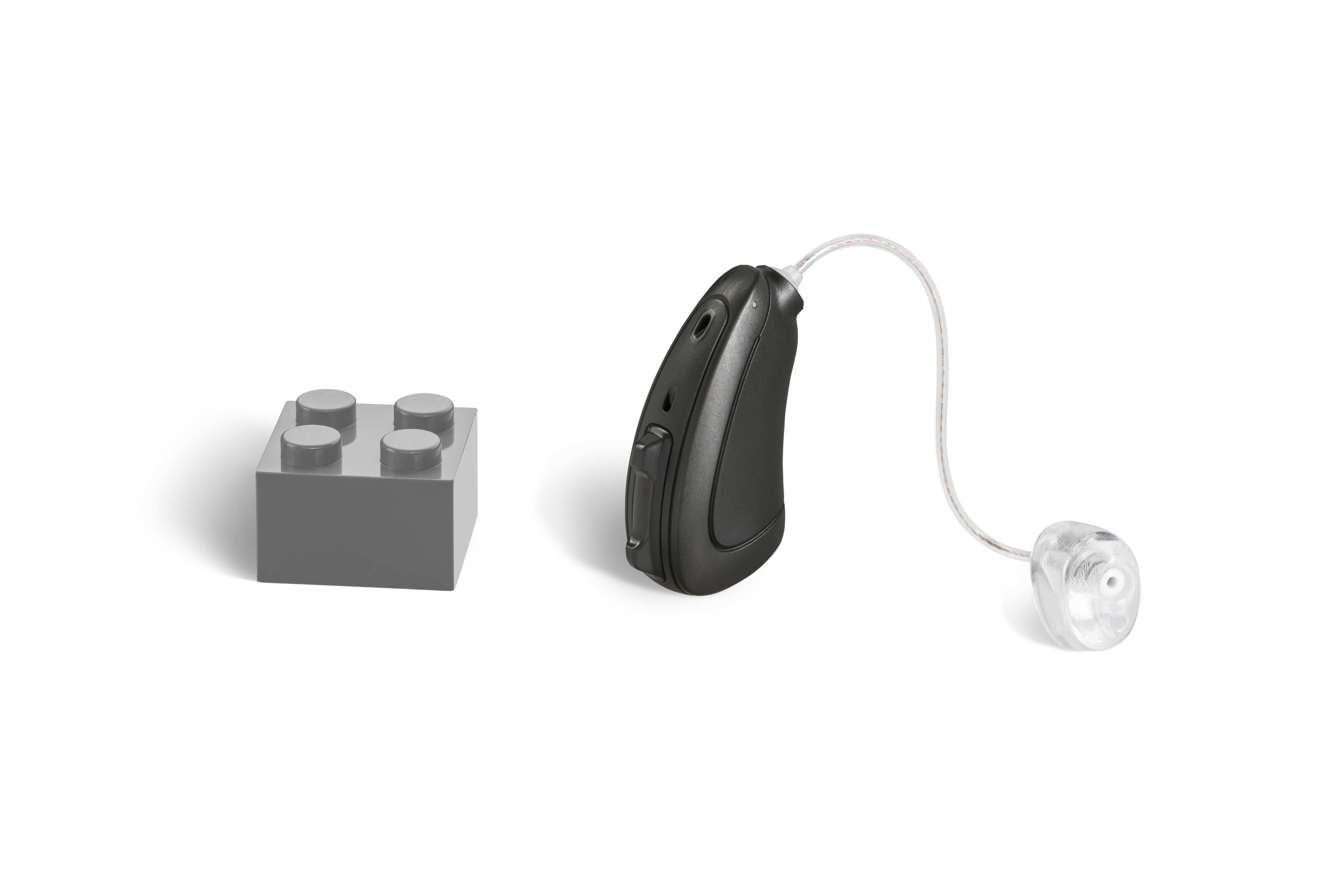 Das Hörgerät KINDwings 2400 R als Bauform HdO mit externem Hörer mit Energieversorgung Akku neben einem grauen Klemmbaustein stehend auf weißem Hintergrund.