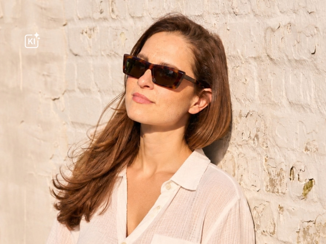Junge Frau mit dezenter Sonnenbrille lehnt an einer Steinwand. 