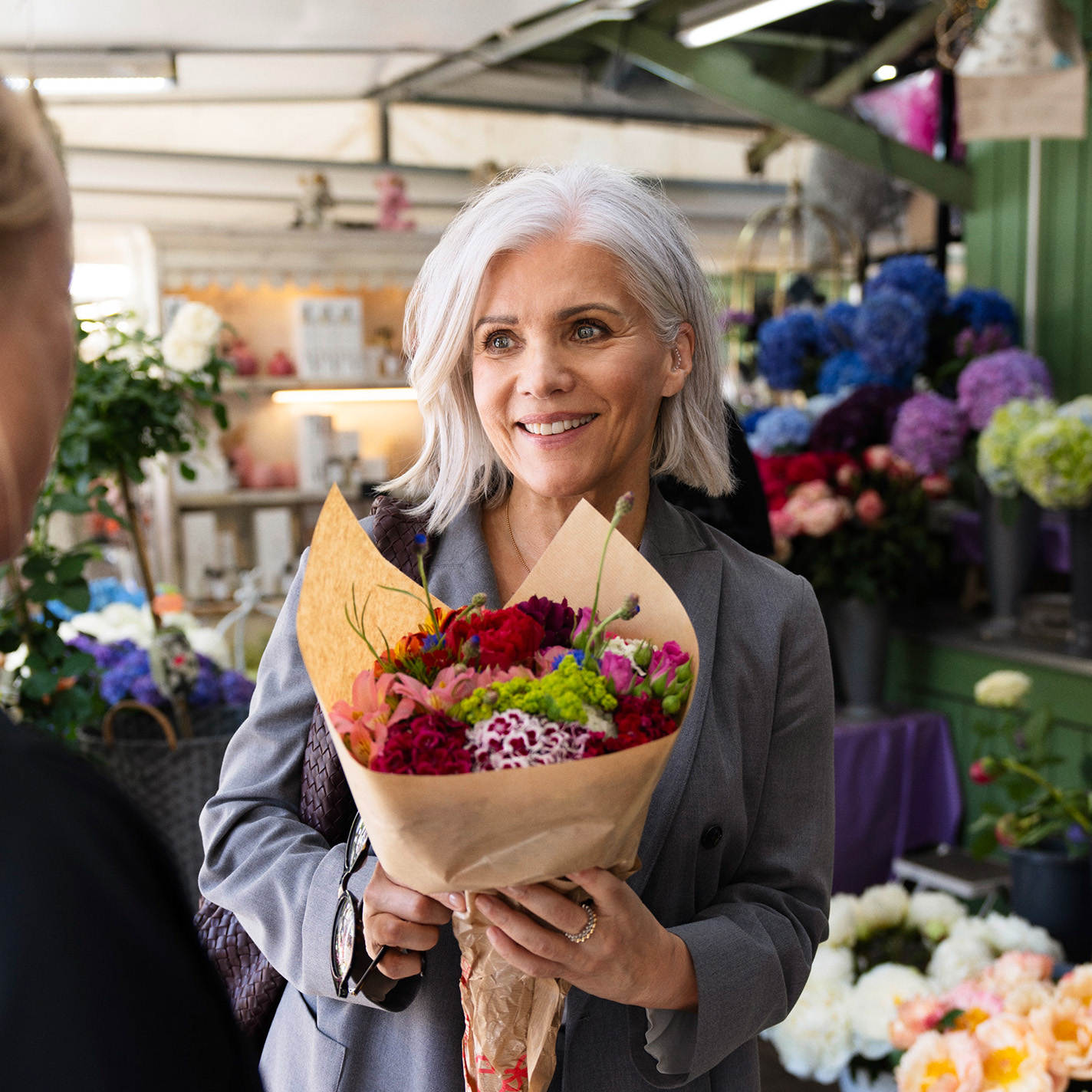 Birgit Schrowange hält einen in Papier eingewickelten Blumenstrauß in der Hand und lächelt eine Verkäuferin an. 