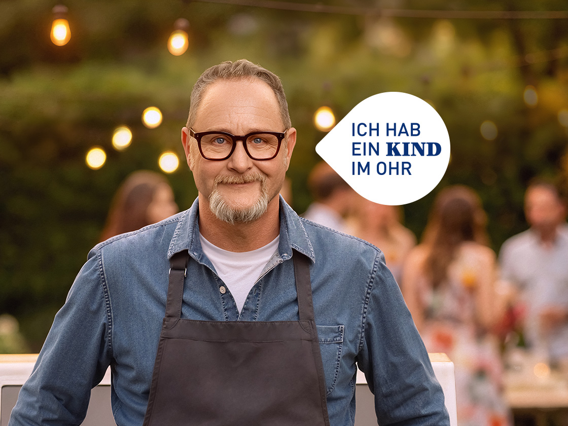 KIND Bekenner Hans-Peter steht mit Grillschürze am Grill, im Hintergrund feiernde Menschen einer Gartenparty.