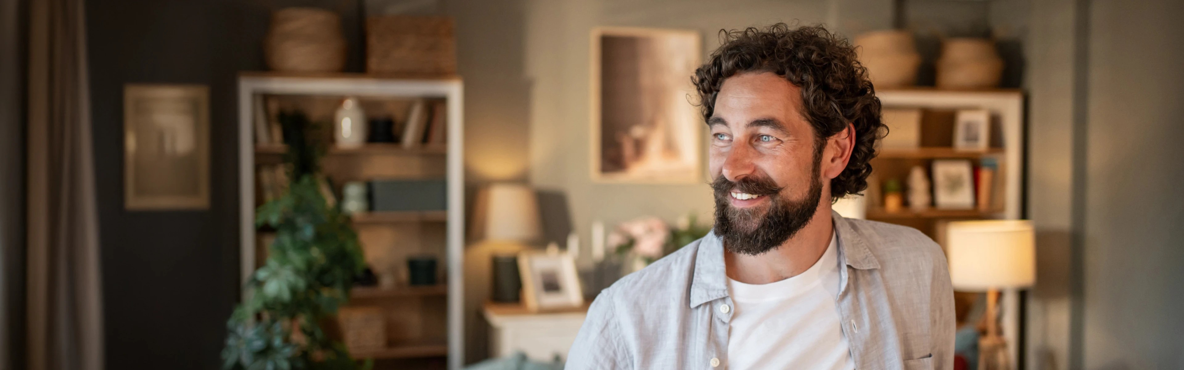 Lächelnder Mann mit dunklen Haaren und Bart steht mit einer Kaffeetasse in der Hand im Wohnzimmer und schaut nach links.
