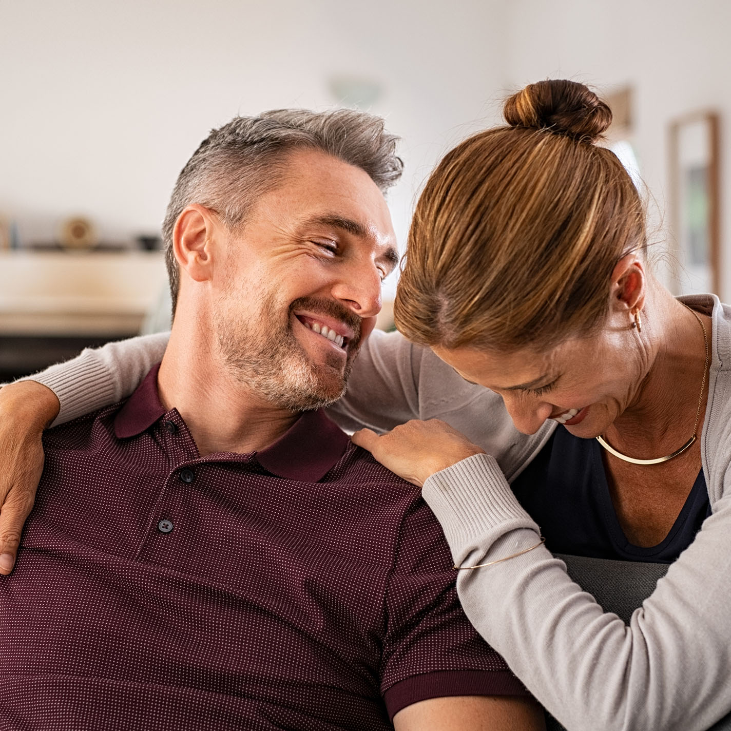 Ein Paar mittleren Alters sitzt im Wohnzimmer, er lehnt seinen Kopf zu Ihr herüber, sie senkt ihren, beide lachen.
