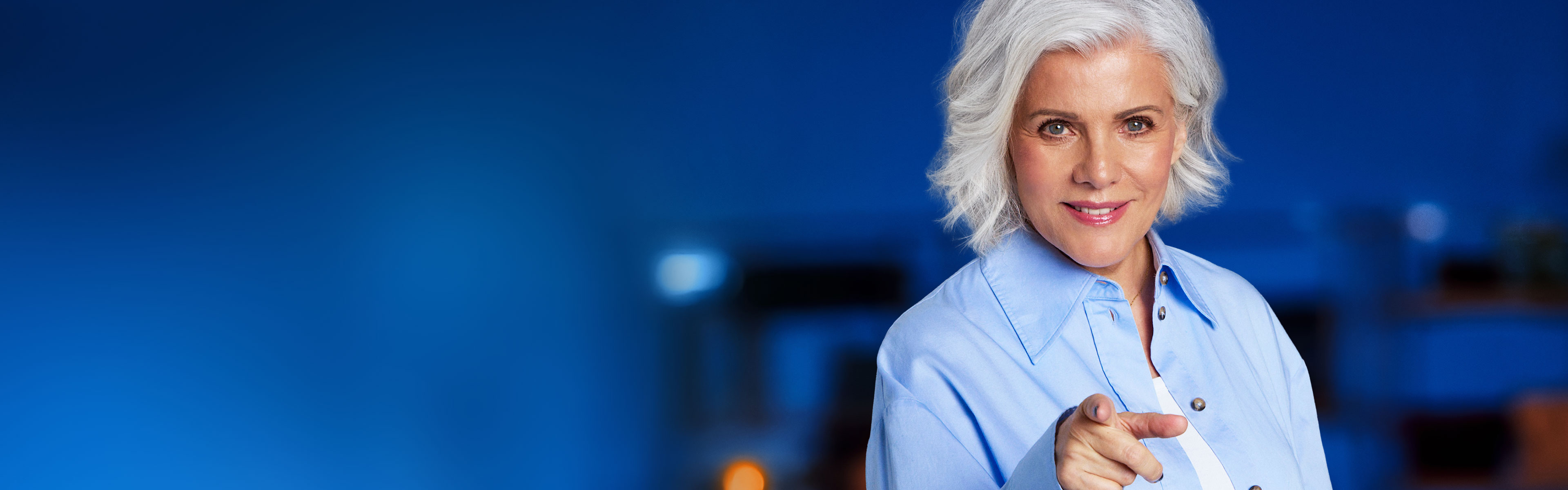 Birgit Schrowange vor blauem, räumlichen Hintergrund, guckt freundlich und zeigt mit dem Finger auf den Betrachter.