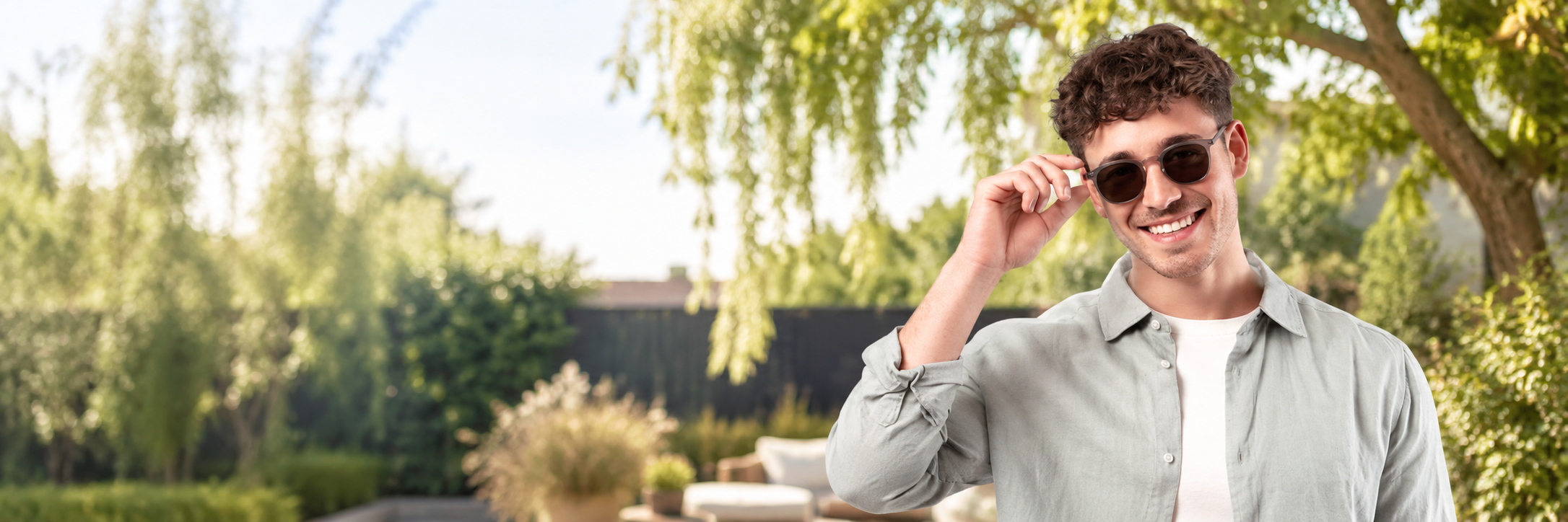 Junger Mann steht im Garten, fässt mit der rechten Hand an seine Sonnenbrille und lächelt.