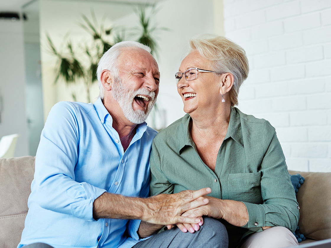 Ein älteres Paar lacht gemeinsam, die Hände ineinander verschränkt, auf der Couch sitzend.